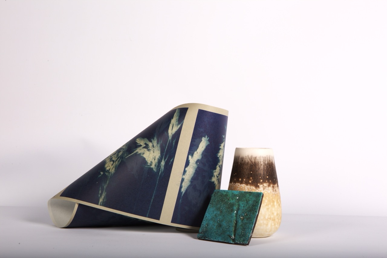 A rolled-up piece of navy-blue paper with a light pattern is placed next to a textured, two-tone ceramic vase, and a small, turquoise square object. The arrangement is set against a plain white background.