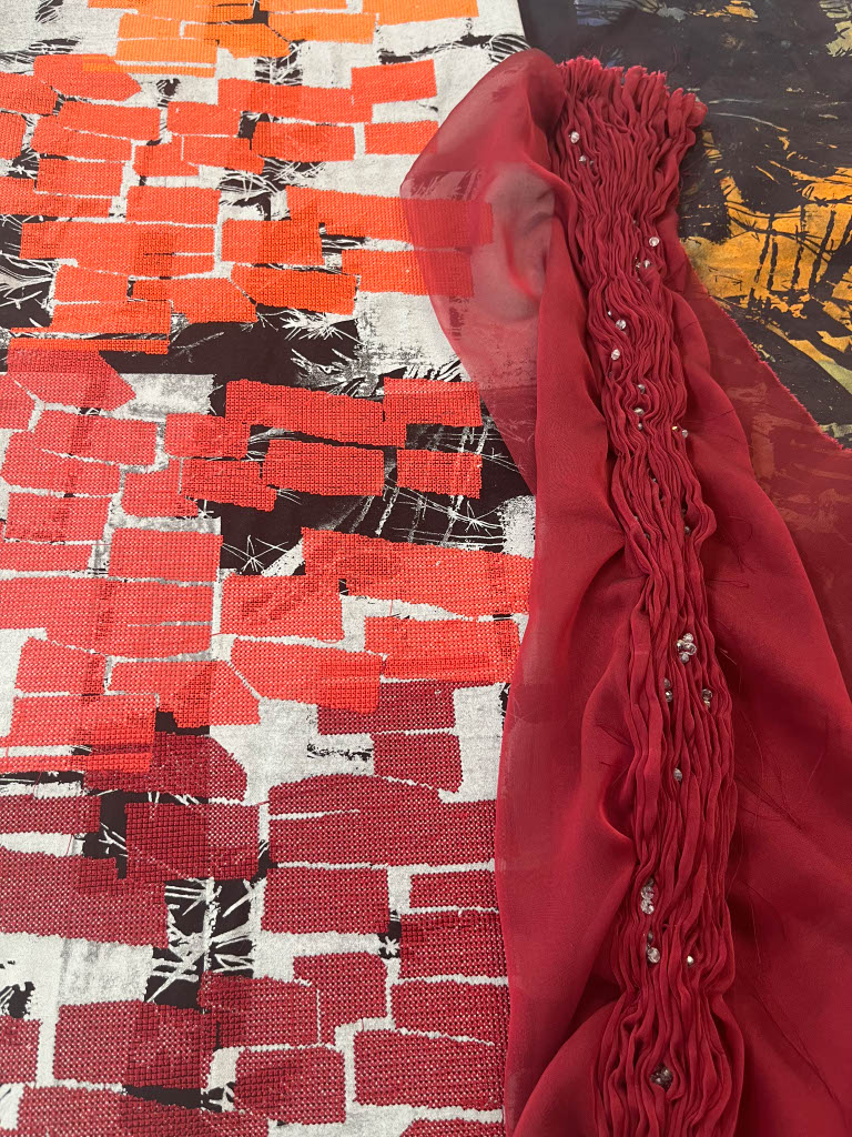 A close-up of two fabrics displayed together on a surface. One fabric features a bold pattern of red and black abstract shapes, while the other is a sheer red fabric with gathered detailing and embellishments along one edge.