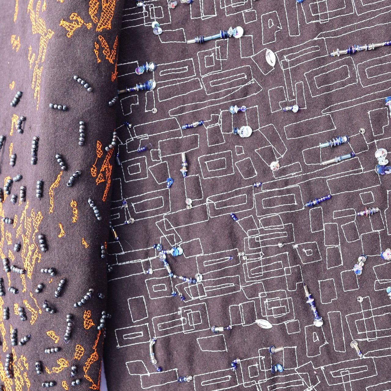 A close-up view of two fabric pieces. One features a vibrant orange design with scattered black beads, while the other displays a light grey geometric pattern embellished with blue and silver beads on a dark background.