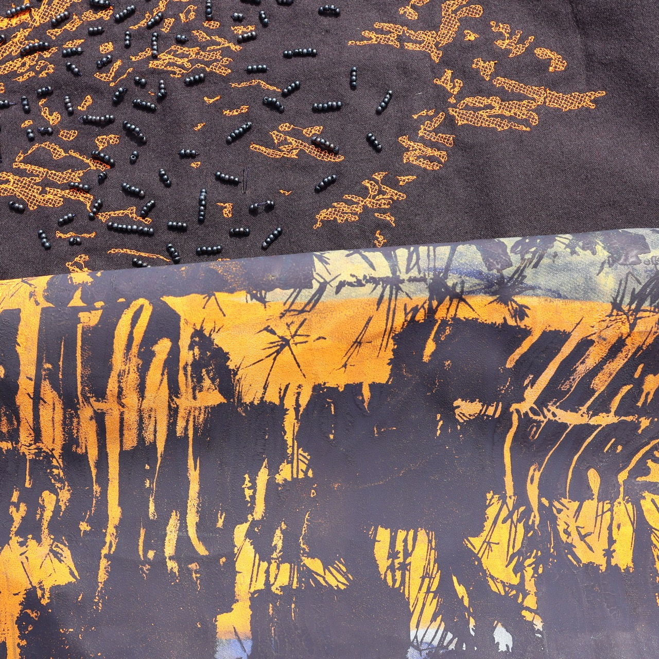 A close-up of two fabric pieces. The top fabric is brown with orange patterns and small black beads arranged in a design. The bottom fabric features an abstract print with orange and grey tones, depicting a bamboo forest under a sunset.