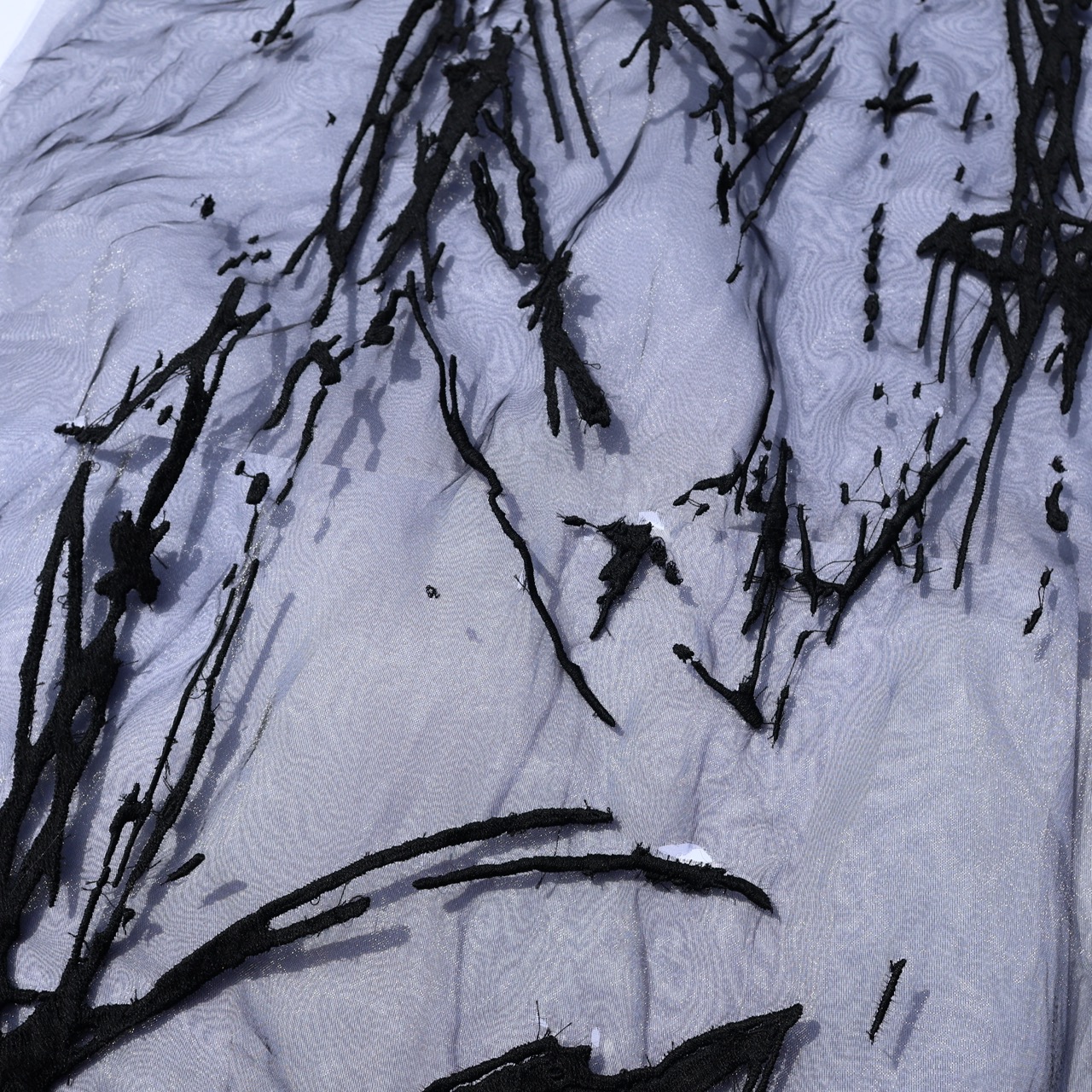 A close-up of a textured fabric featuring black, abstract brushstroke patterns on a grey background. The design consists of elongated, tangled lines resembling twigs or branches, creating a layered visual effect.