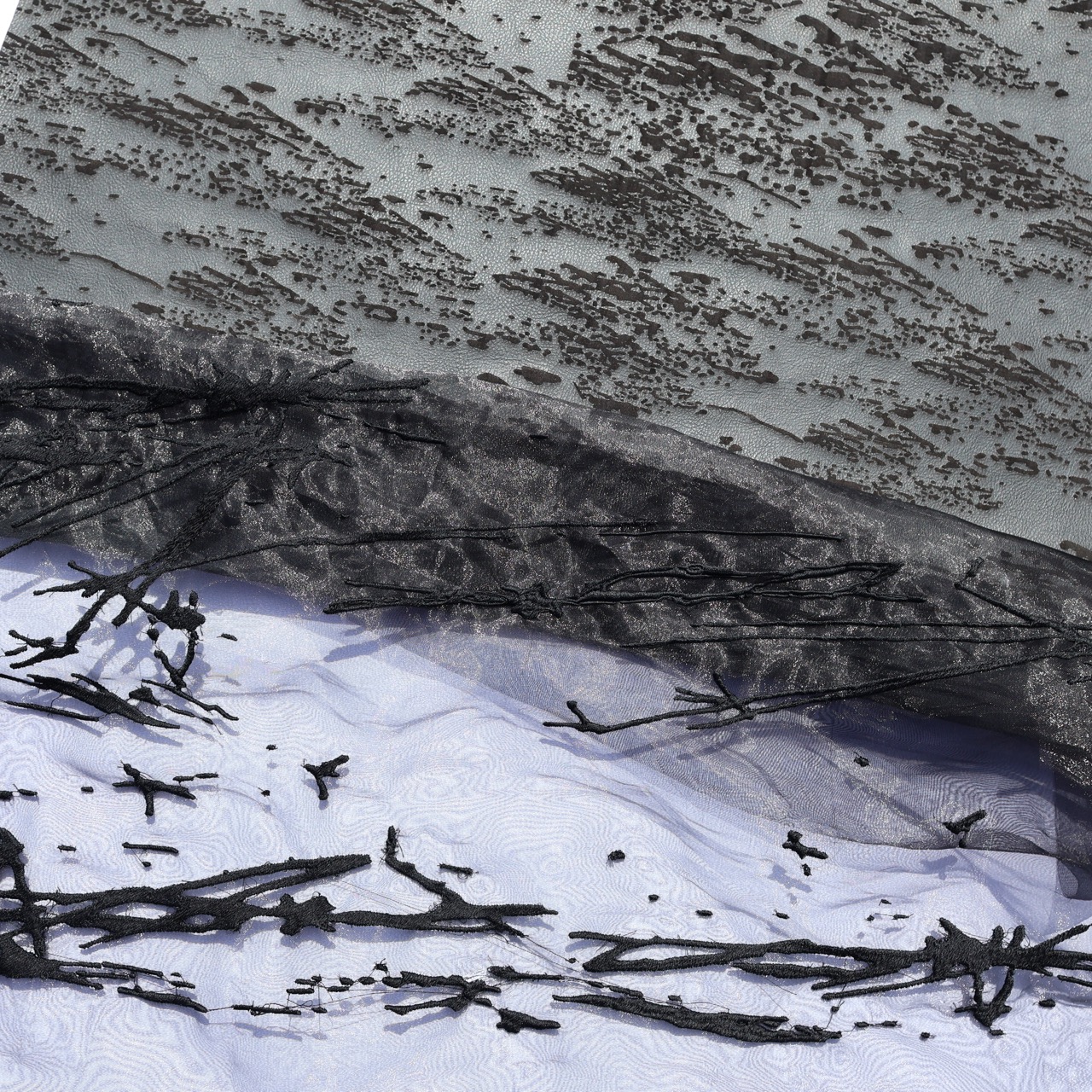 A textured fabric featuring a grey background with a pattern of small black dots, layered over a sheer black material. Below, intricate black thread forms abstract shapes against a light, patterned surface.