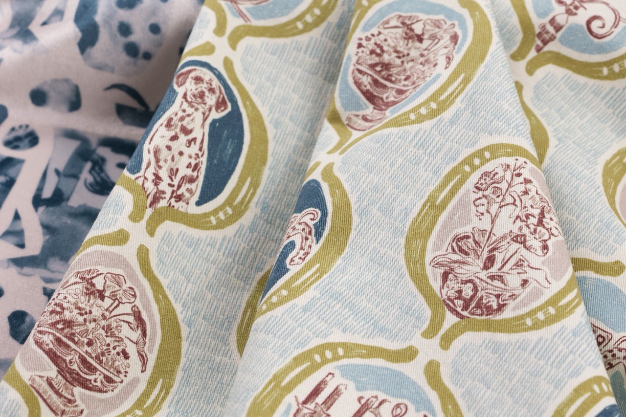 Close-up of two pieces of fabric featuring decorative patterns. One fabric displays blue and green motifs with red and pink floral designs inside oval shapes, while the other has a predominantly light pink background with darker blue abstract patterns.