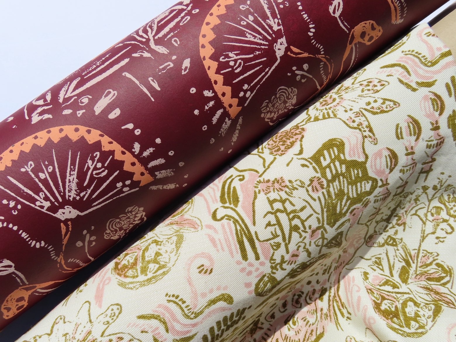 Two rolls of patterned fabric laid side by side. The top roll features a deep red background with floral and fan designs in a lighter tone, while the bottom roll has a cream background with green and pink botanical illustrations.