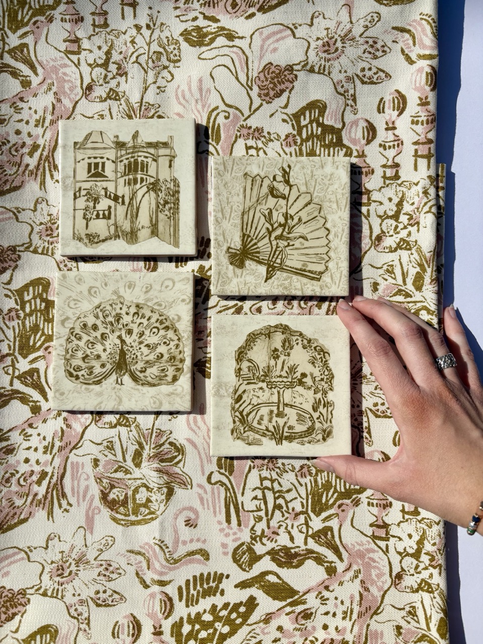 Four decorative ceramic tiles arranged on a patterned fabric background. The tiles feature various designs including a peacock, flowers, and a house, while a hand with a ring is visible touching one tile.