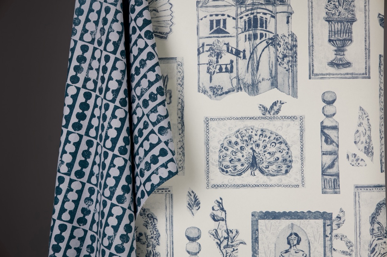 A patterned fabric hanging against a wall featuring a blue design of peacocks, classical motifs, and floral illustrations on a white background. A second fabric with a different pattern in shades of teal is draped over the edge, showcasing circular designs.