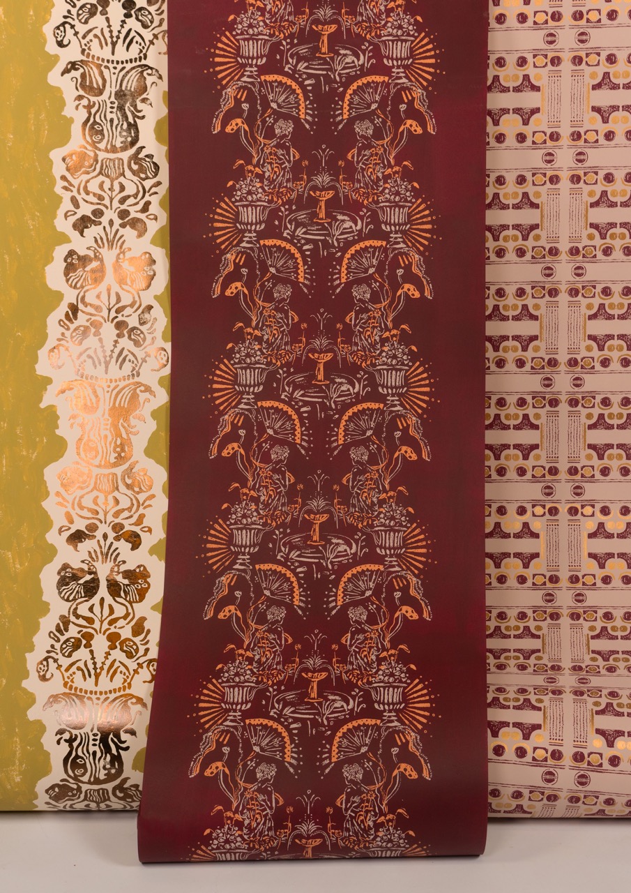 Three rolls of patterned wallpaper displayed together. The left roll features a green background with intricate white designs, the middle roll is burgundy with orange motifs of plants and figures, and the right roll has a patterned design in pale purple and cream.