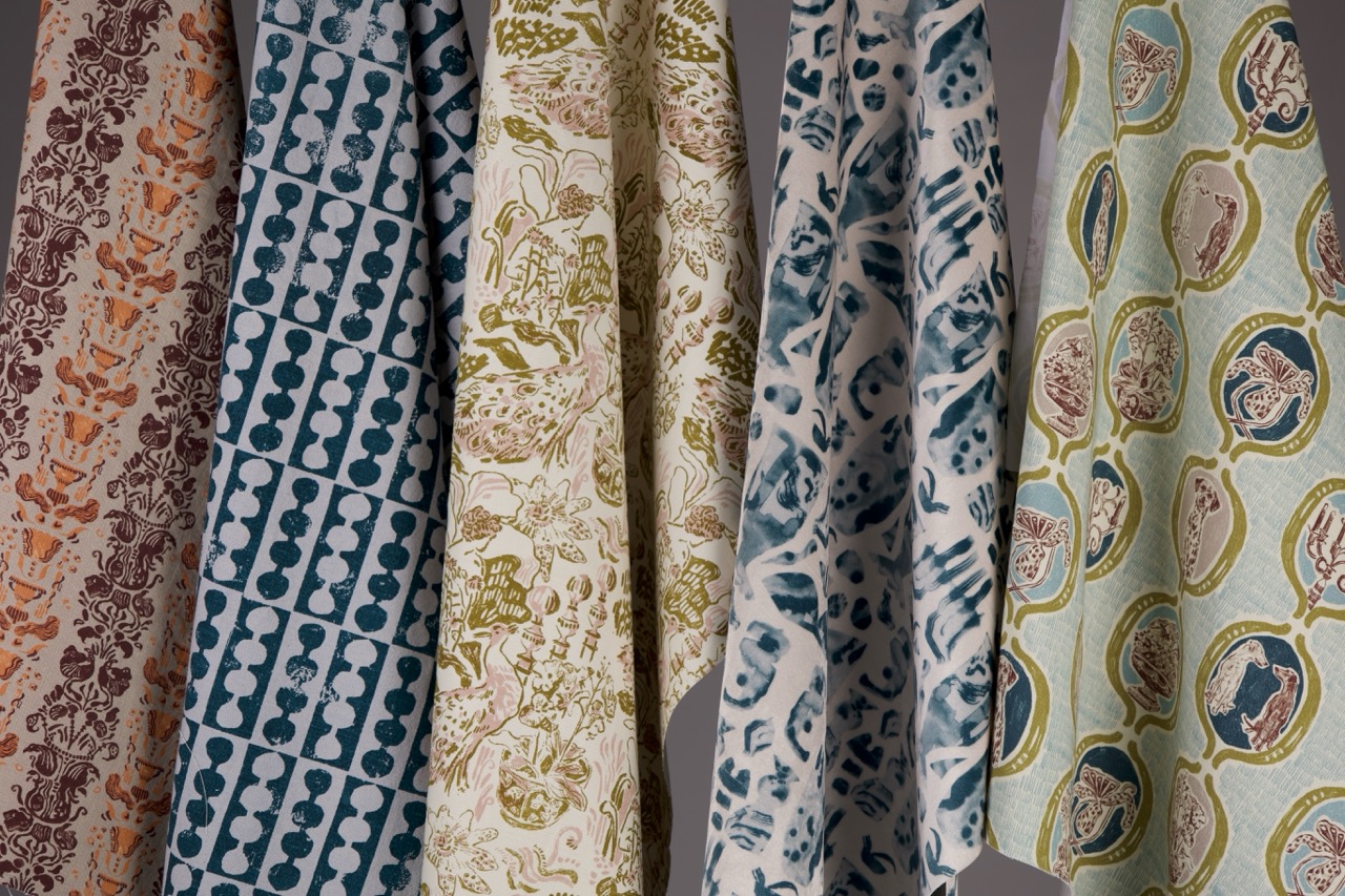 A collection of six patterned fabrics hanging side by side. The designs include floral motifs, geometric shapes, and paisley patterns in various colours such as blue, green, brown, and cream against a neutral background.