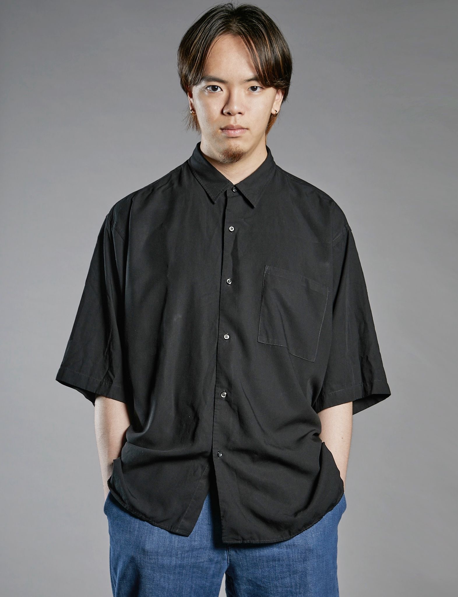 A young man with brown hair, wearing a black short-sleeved shirt with a front pocket, stands facing the camera. He has his hands in his pockets and is wearing blue trousers, with a neutral grey background behind him.