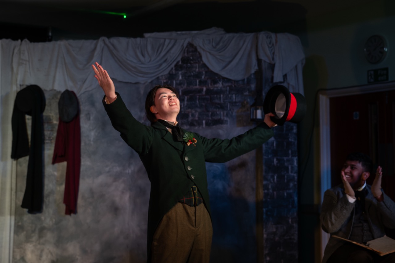 A young performer in a black and green costume holds a top hat aloft with both arms raised, standing on a stage. The background features draped fabric and costumes hanging on a wall, while an audience member is seated in the foreground, applauding.