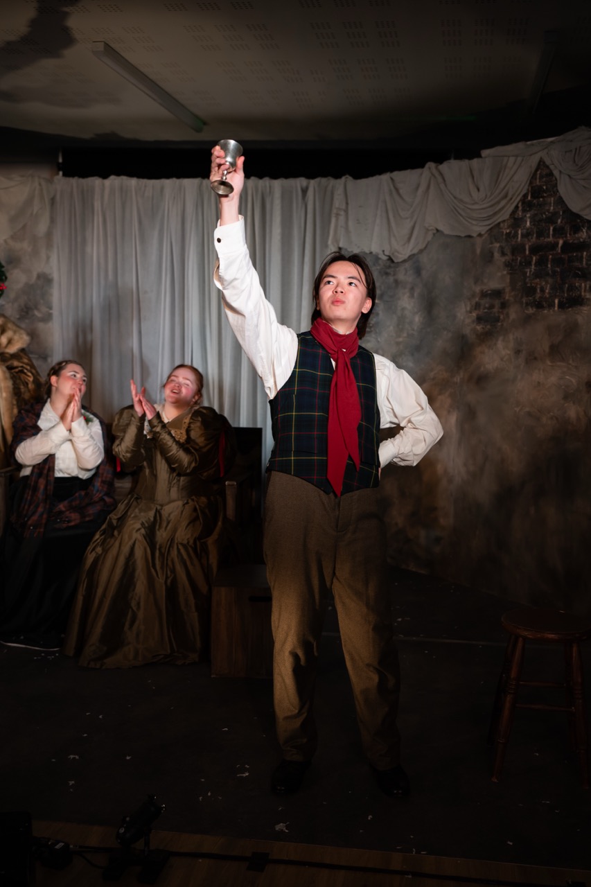 A young performer in period costume holds a cup aloft with one arm raised, standing confidently on a stage. Behind, three audience members in historical attire watch attentively, with two women seated and one clapping in appreciation.