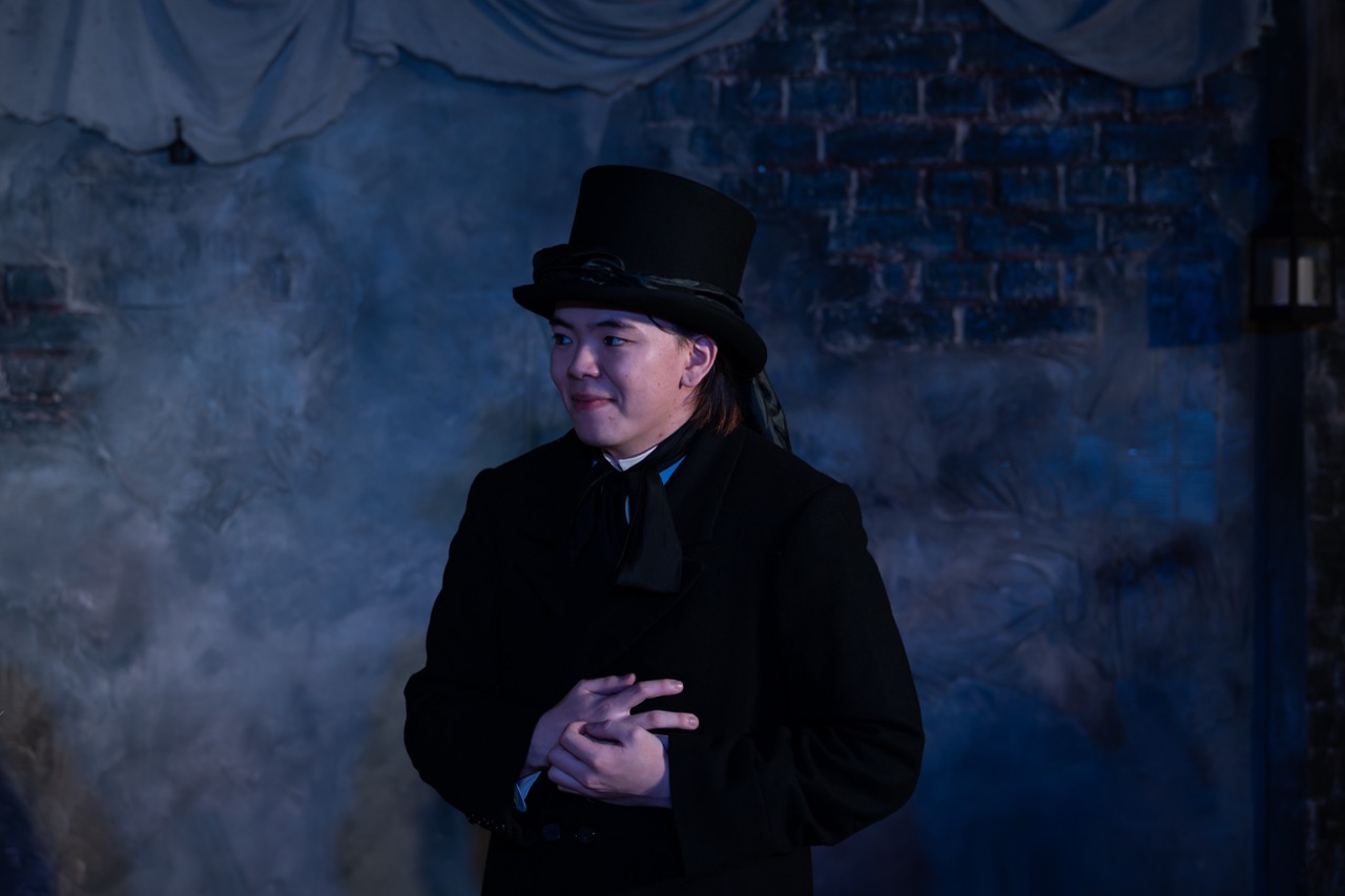A person dressed in a black coat and top hat stands with their hands clasped in front of them. The background features a smoky grey texture and brick wall, creating a dramatic atmosphere.