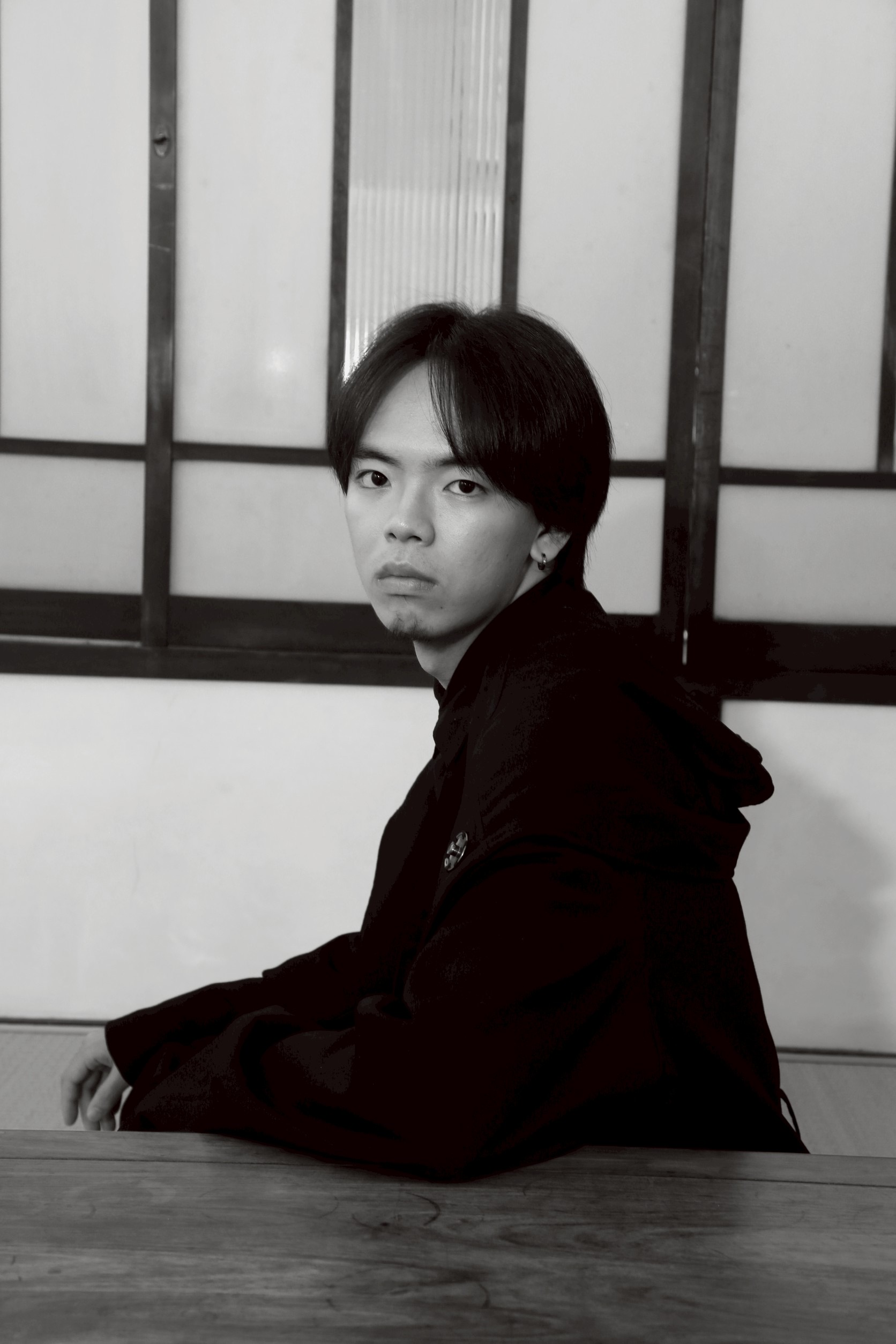 A young man with short black hair is seated at a wooden table, looking towards the camera. He is wearing a black hoodie and the background features a wall with large glass panels.