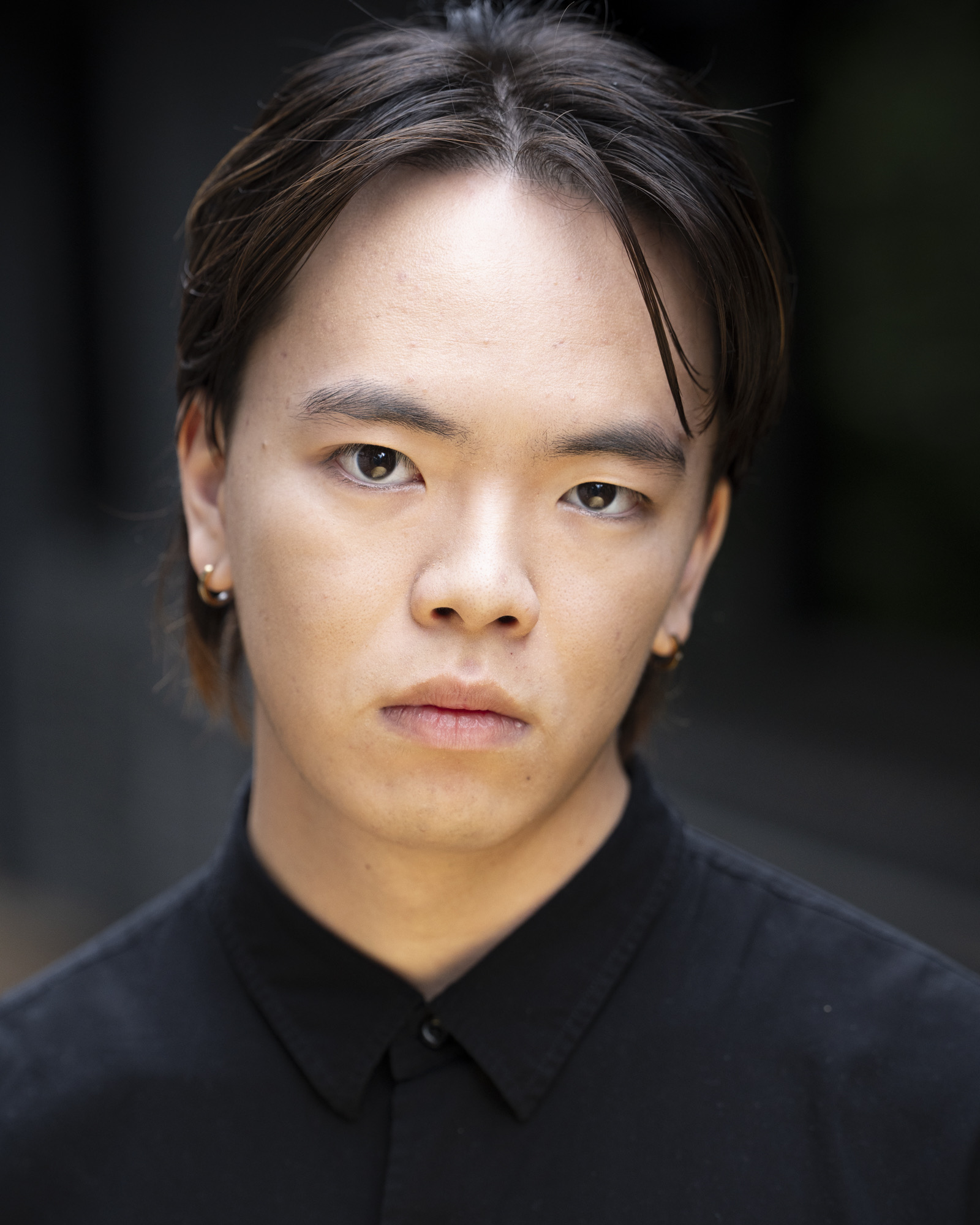 A young man with straight black hair and wearing a black shirt is posed in front of a blurred background. He has earrings and is looking directly at the camera, highlighting his facial features.