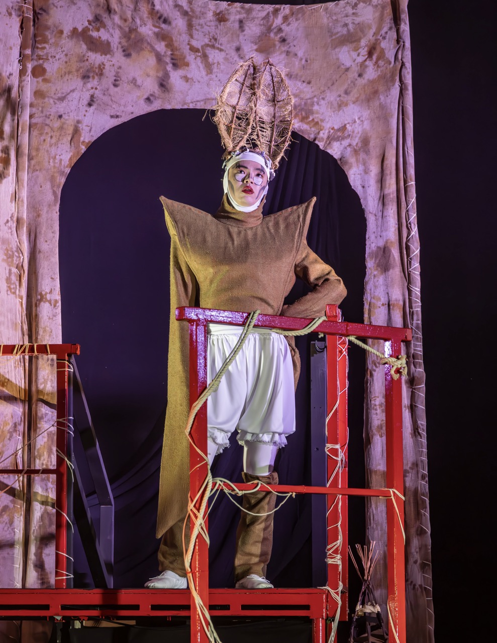 A performer wearing a brown costume with a structured design stands on a red wooden platform. The backdrop features a dark curtain and textured fabric, highlighting the theatrical setting.