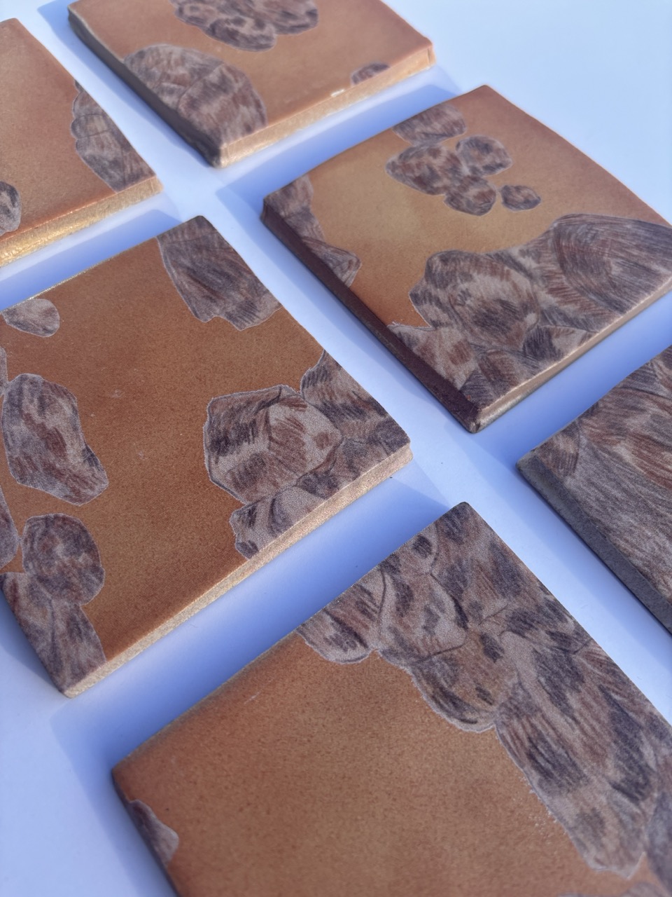 Six handmade tiles displayed on a light blue surface. Each tile features a brown and orange design with abstract shapes resembling rocks, showcasing a textured appearance.