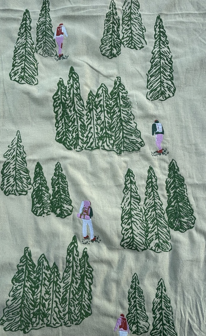 A patterned fabric featuring green trees and small human figures. The figures, in varying colours, appear to have backpacks and are depicted amongst tall, stylised green coniferous trees on a light background.