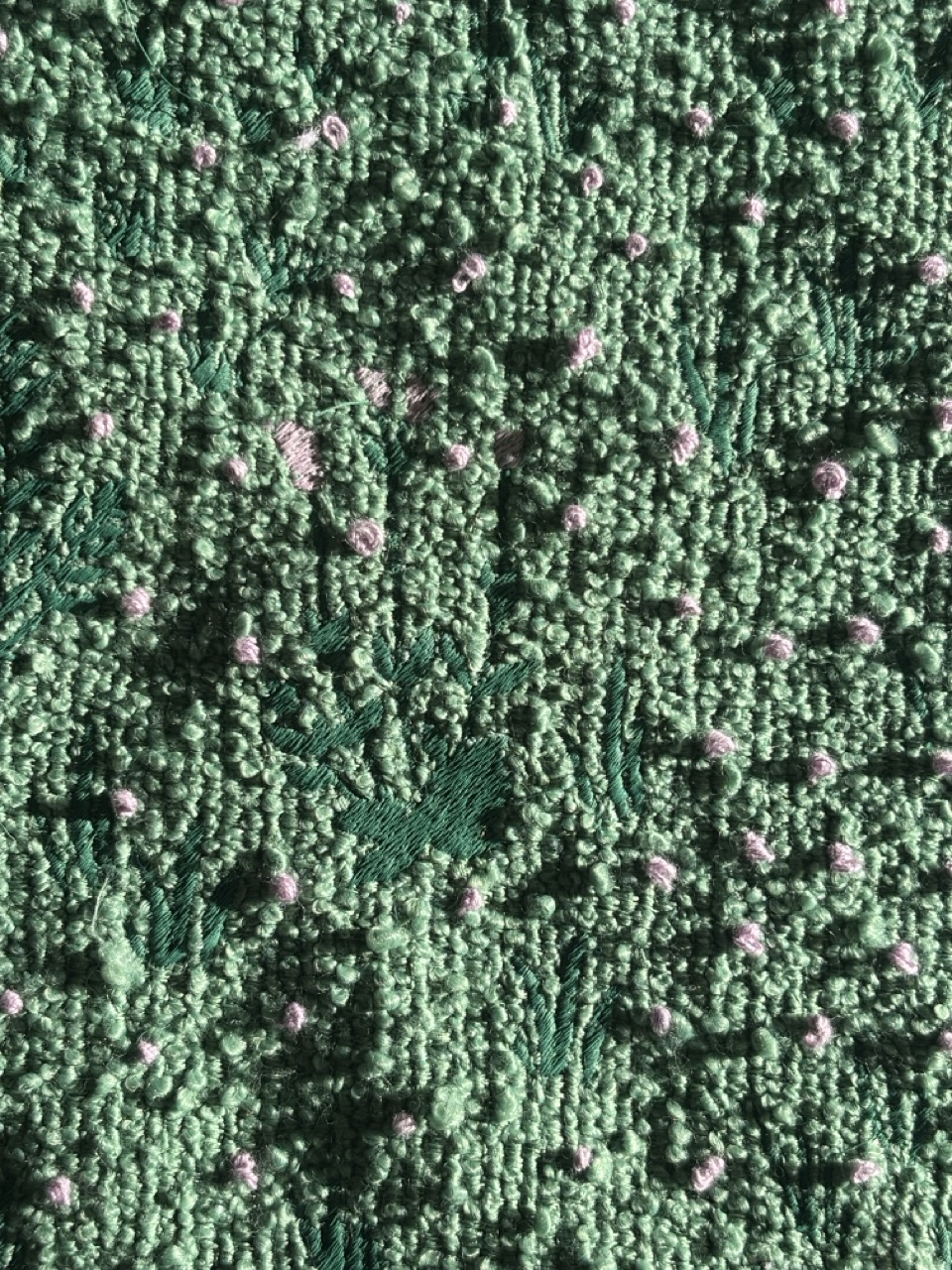 A textured green fabric featuring a pattern of raised bumps and floral designs in darker green and light pink accents. The fabric has a soft, textured surface and is illuminated by natural light.