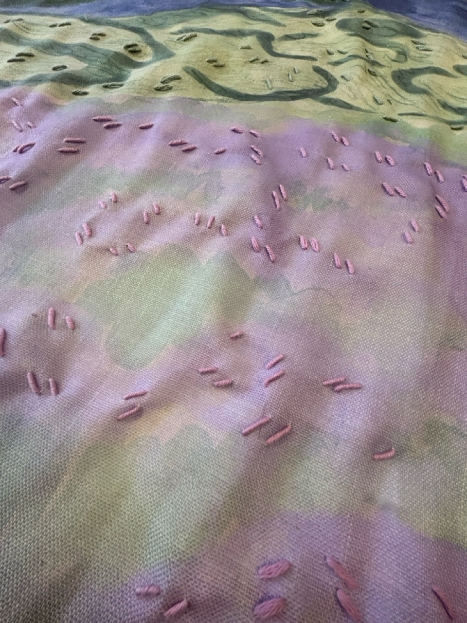 A close-up view of a fabric with a textured surface, featuring various shades of pink and green hues. Small embroidered shapes in pink are scattered across the surface, creating a tactile pattern on the woven material.