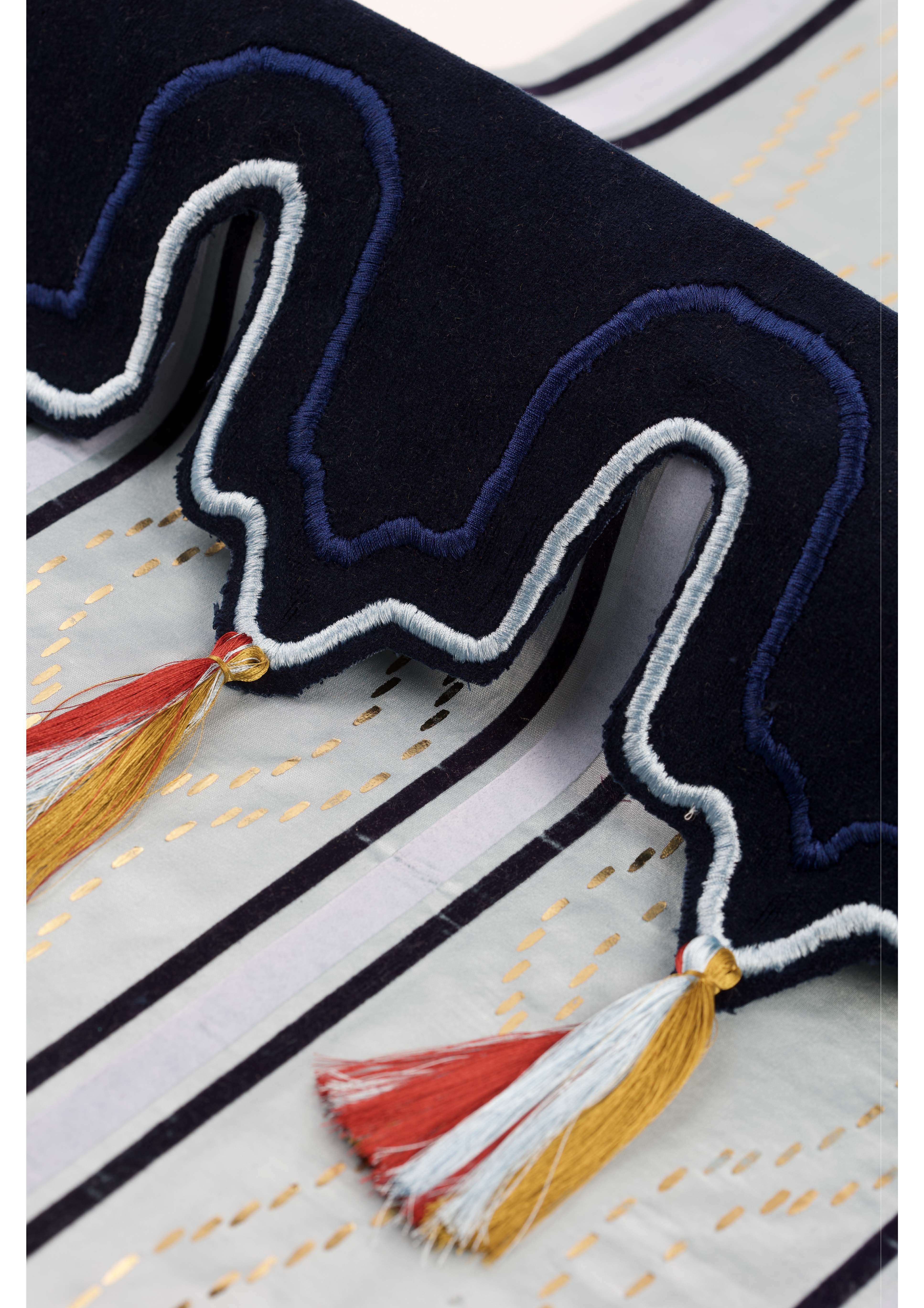 A close-up of a decorative fabric with a navy blue wavy border featuring white embroidery. The border has three tassels in red, yellow, and white, suspended from it, with a patterned, light background displaying thin, dark stripes.