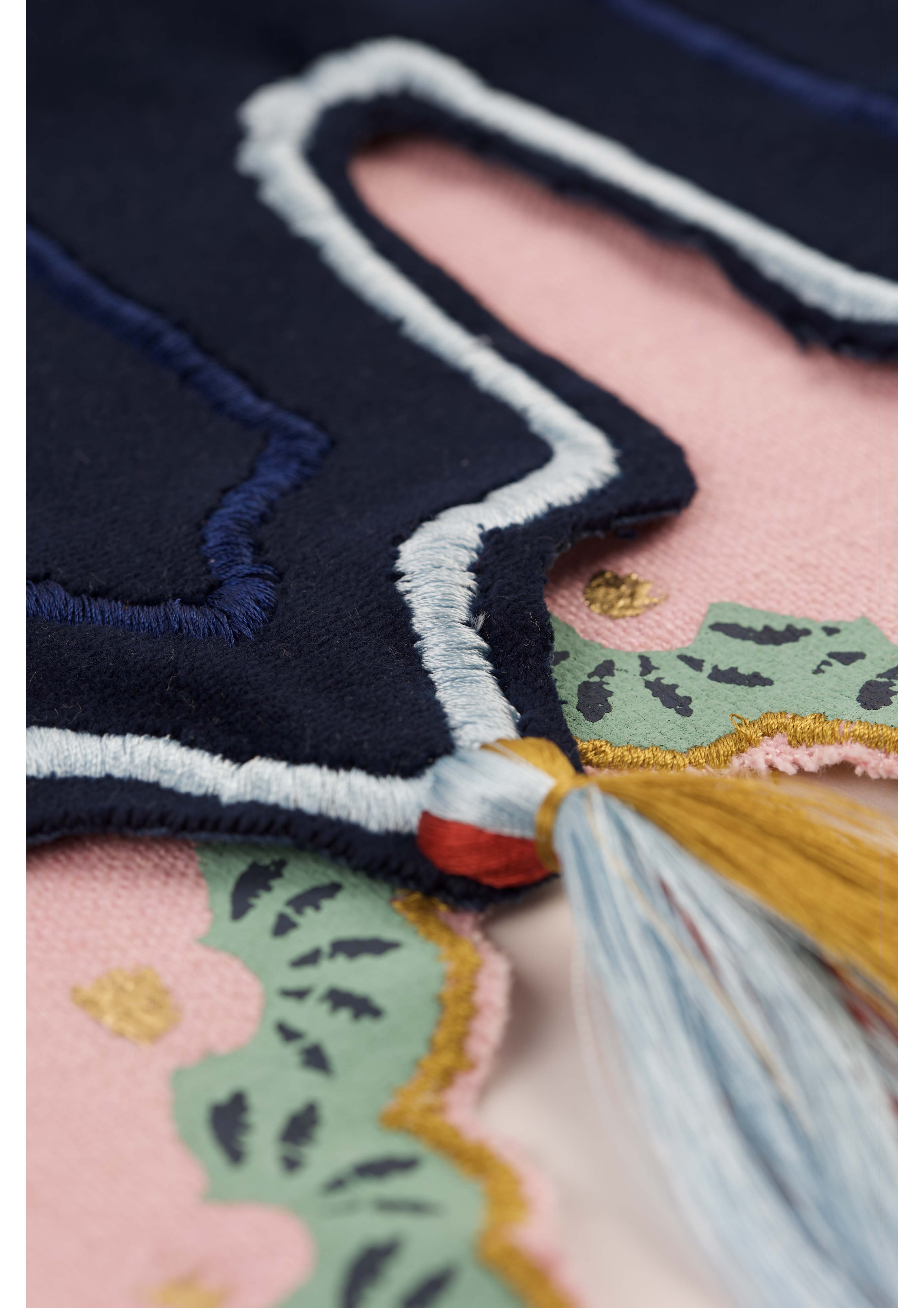 Close-up of a navy blue fabric with embroidered detailing, featuring a patterned trim in green and pink. A decorative tassel in blue, yellow, and red hangs from the edge, adding a textured element to the design.