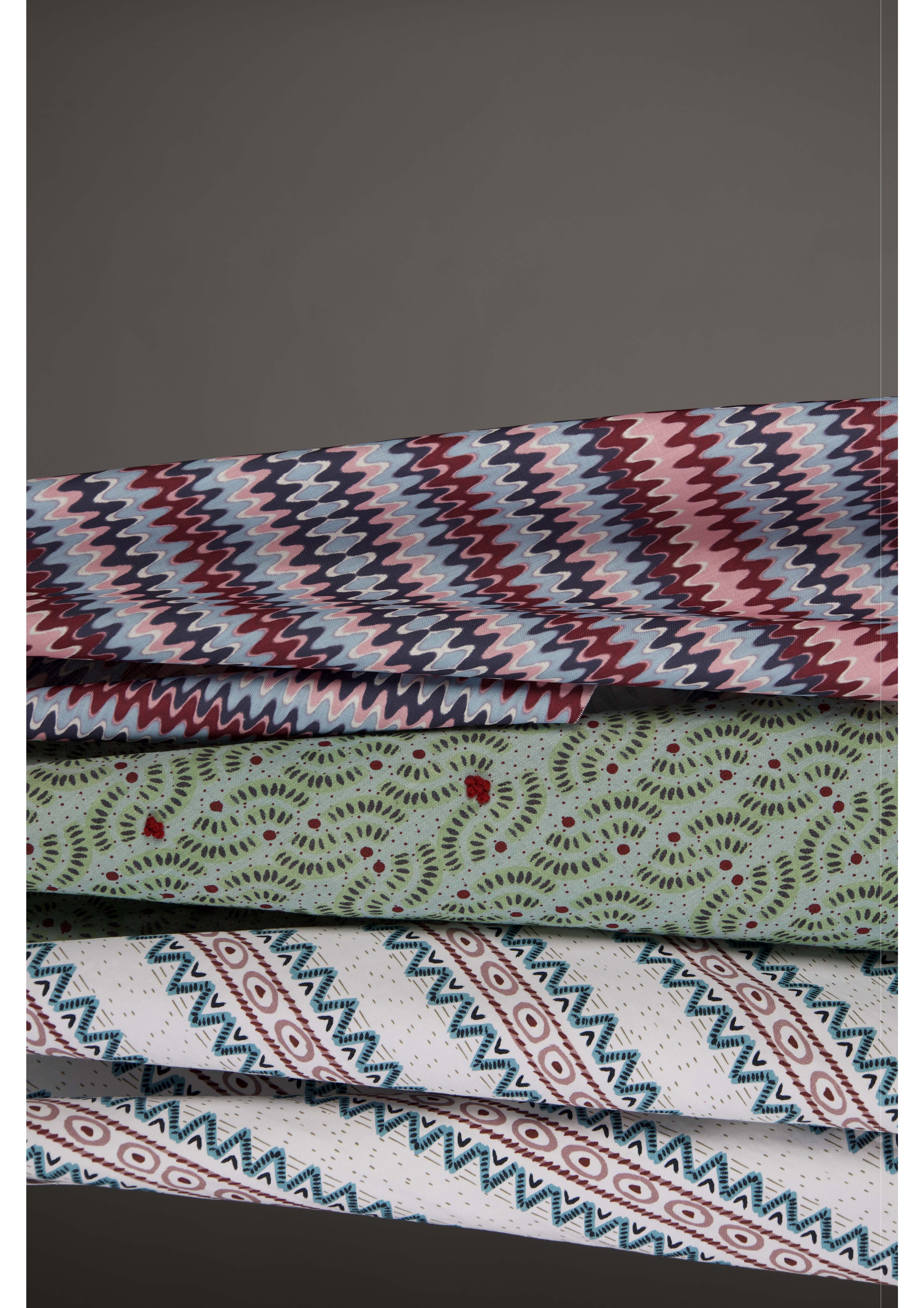 Four folded pieces of patterned fabric stacked on top of each other. The top fabric features a zigzag pattern in pink, red, and blue. Below it is a green fabric with a circular design in red. The third fabric is white with a blue geometric pattern, and the bottom fabric has a floral motif.