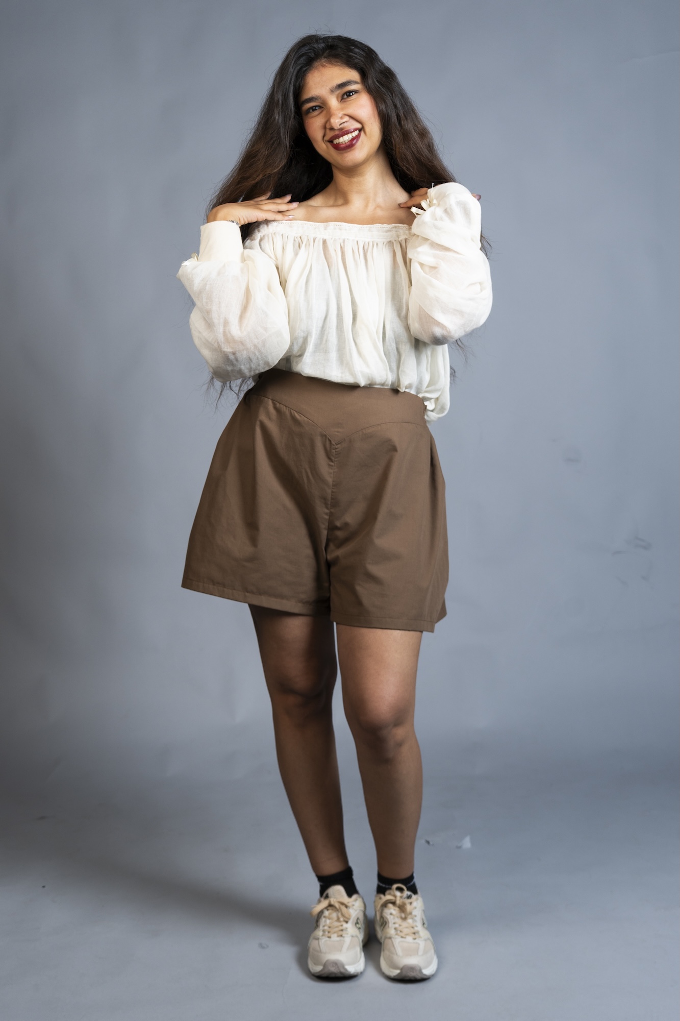 A model wears a white off-shoulder blouse, brown shorts and beige trainers against a plain grey studio backdrop.