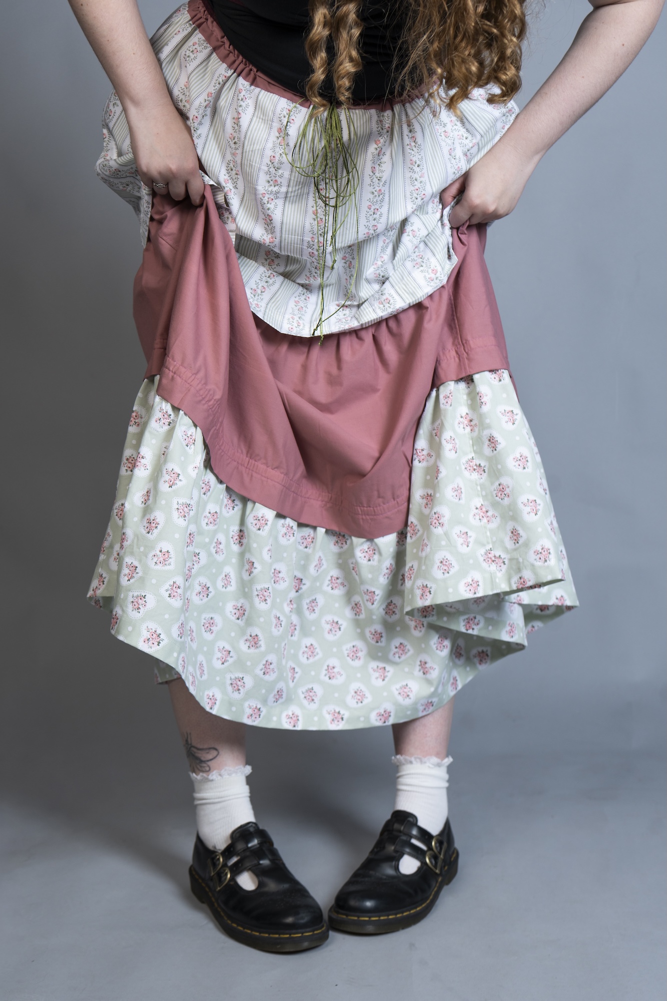 A model wearing a layered pink and floral dress with black shoes and white socks, standing against a plain grey background.