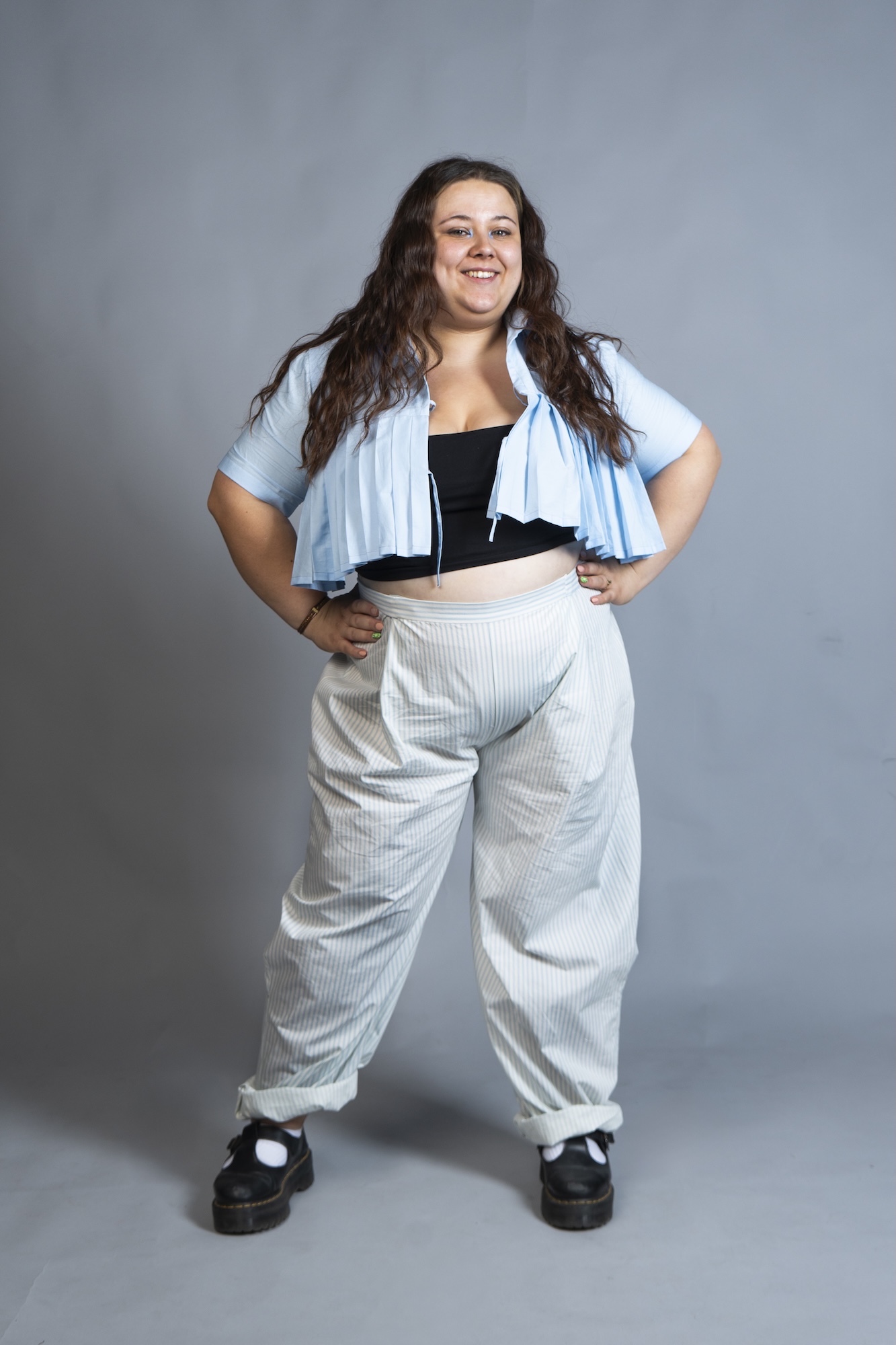 A model with long wavy hair stands with hands on hips, wearing a light blue cropped top, white trousers and black shoes against a plain grey backdrop.
