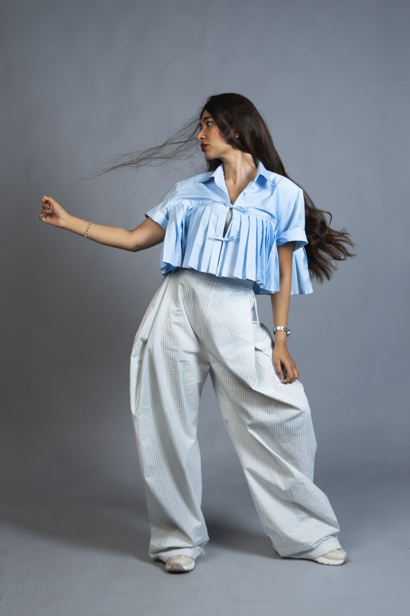 A model wearing a light blue ruffled shirt and wide white trousers stands against a grey studio backdrop, with one arm extended and long hair blowing sideways.