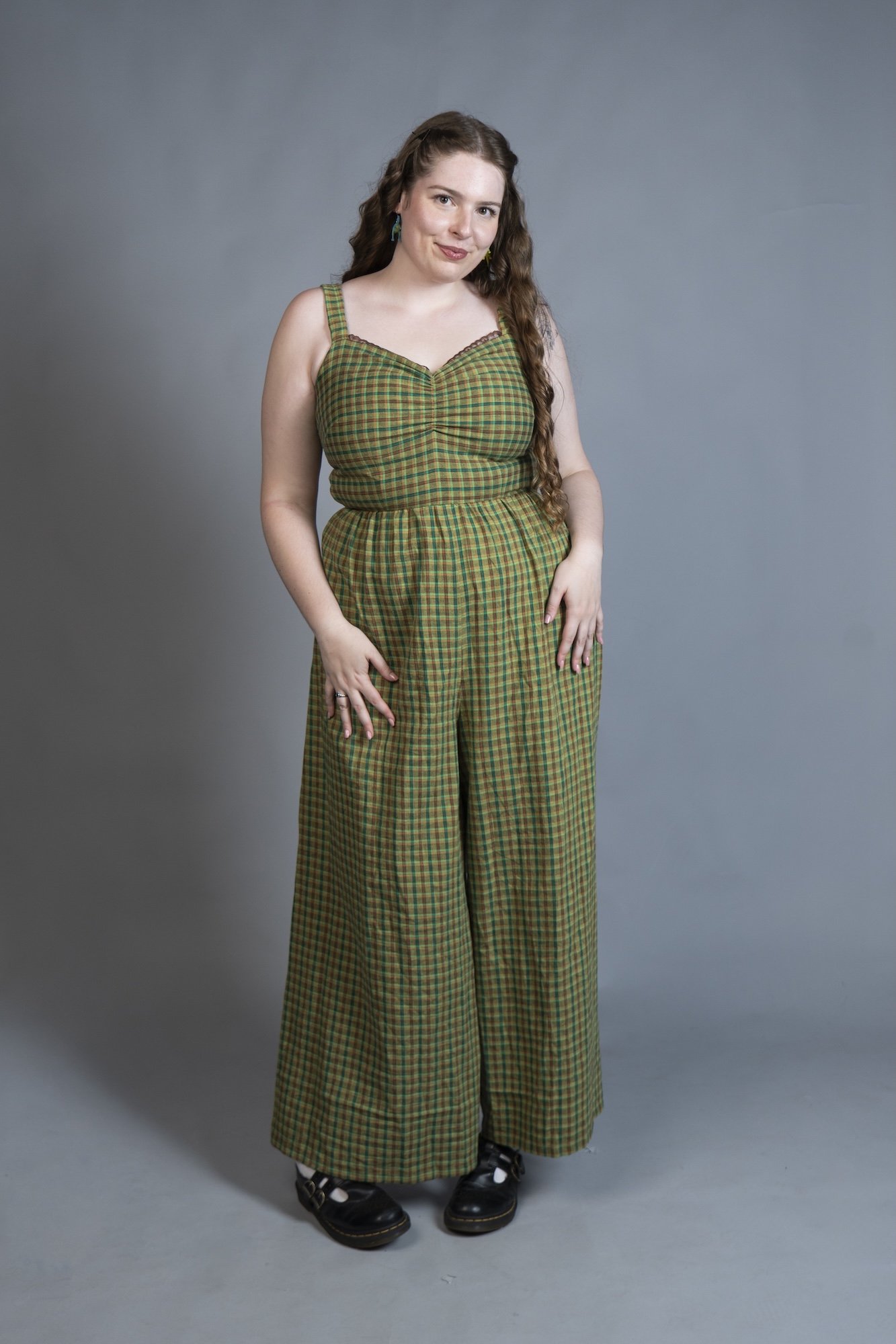 A model in a green checked jumpsuit stands against a plain grey studio backdrop, wearing black shoes.