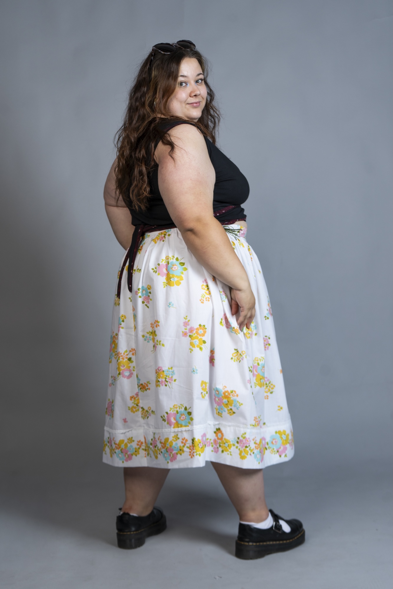 A model wears a black top and a white floral skirt, standing against a plain grey studio background.