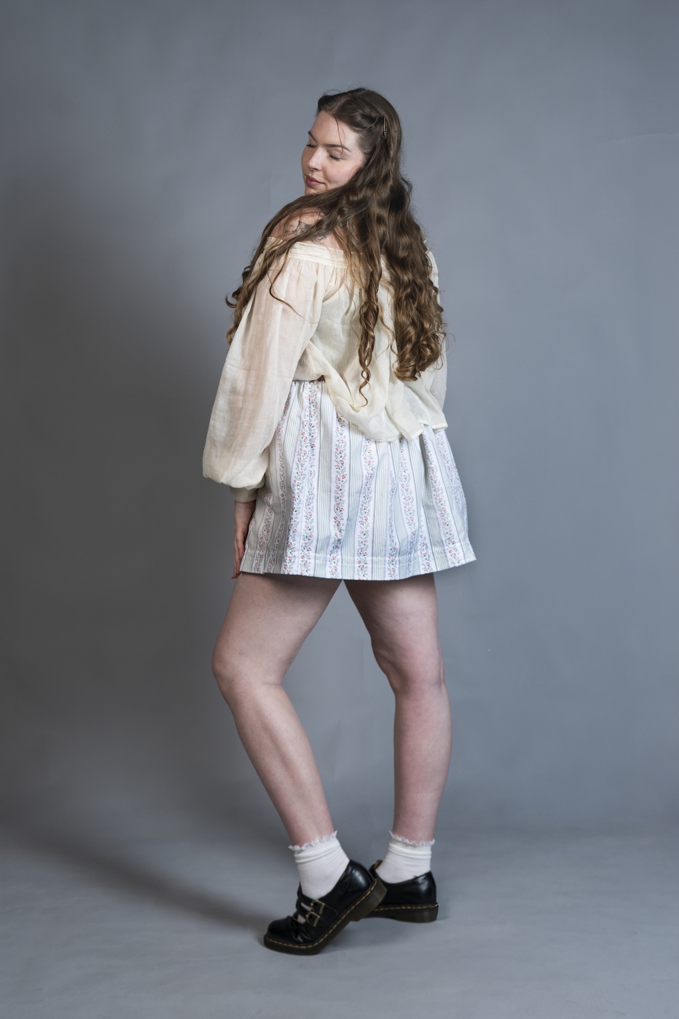 A model wears a cream blouse and striped skirt, standing in black shoes against a plain grey studio backdrop.