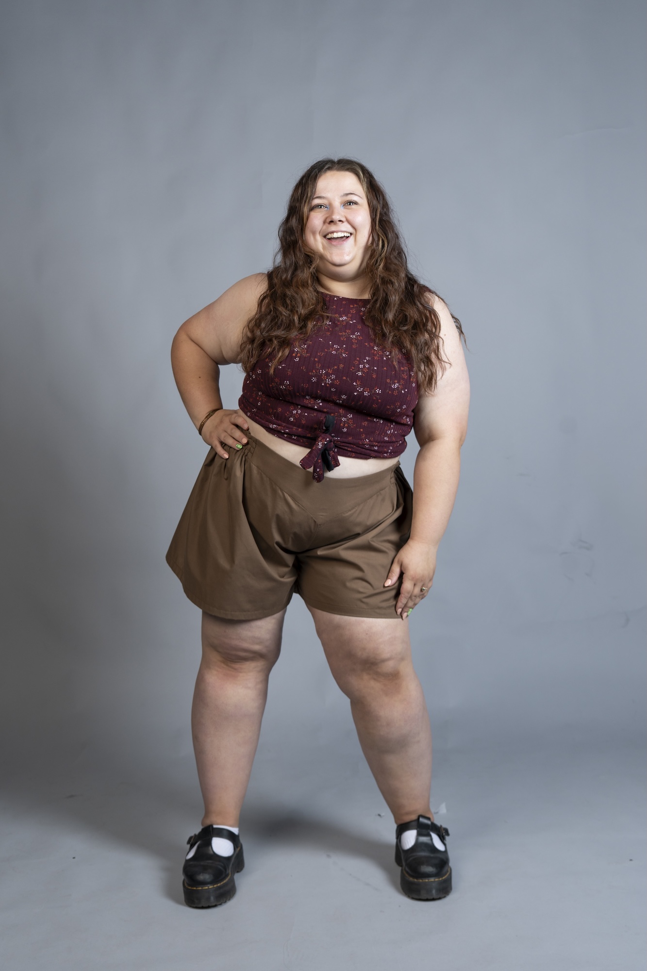 A model stands posing against a plain grey backdrop, wearing a maroon sleeveless top, brown shorts and black shoes.