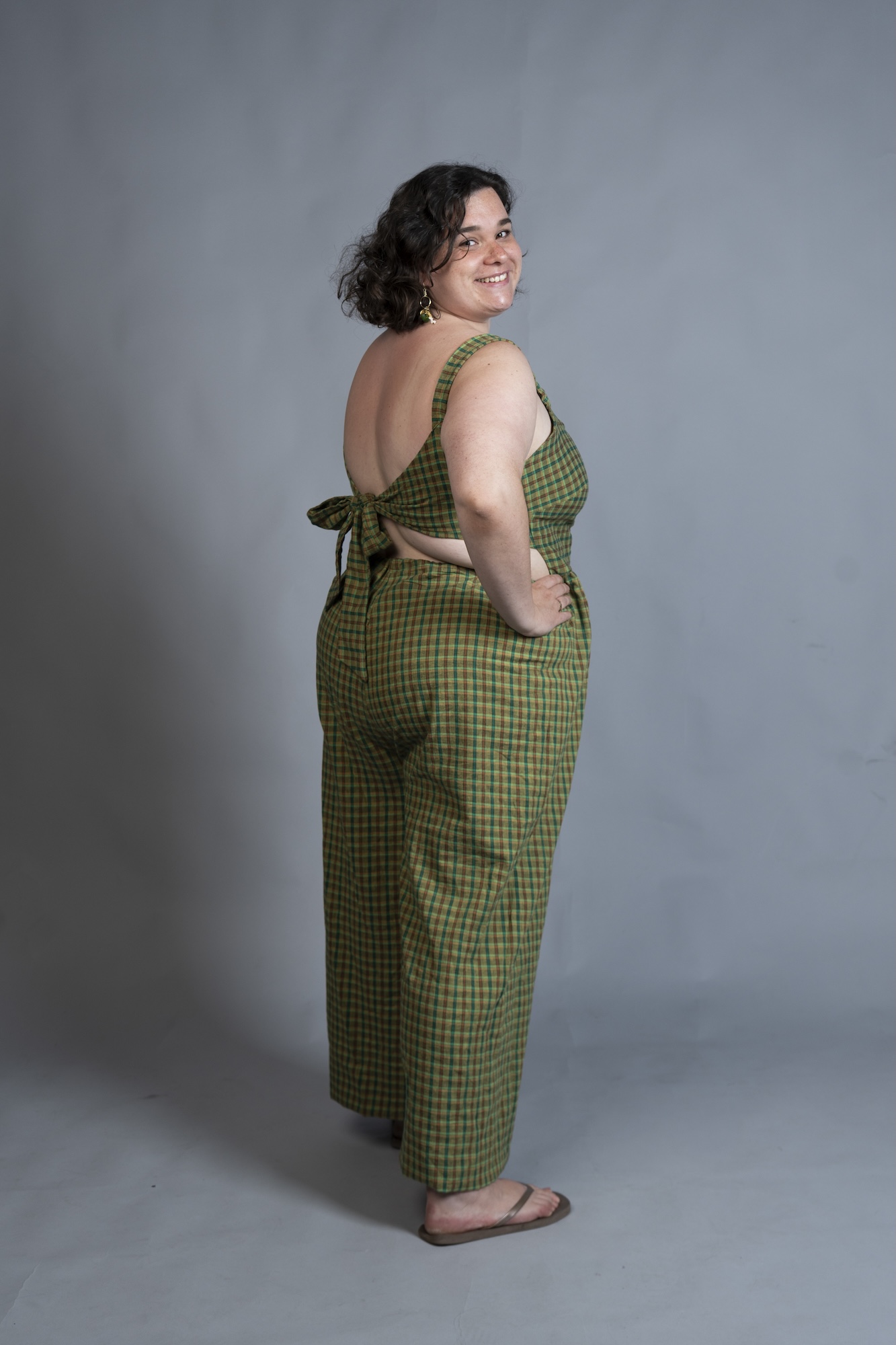 A model in a green checked sleeveless jumpsuit stands in profile against a plain grey studio background.