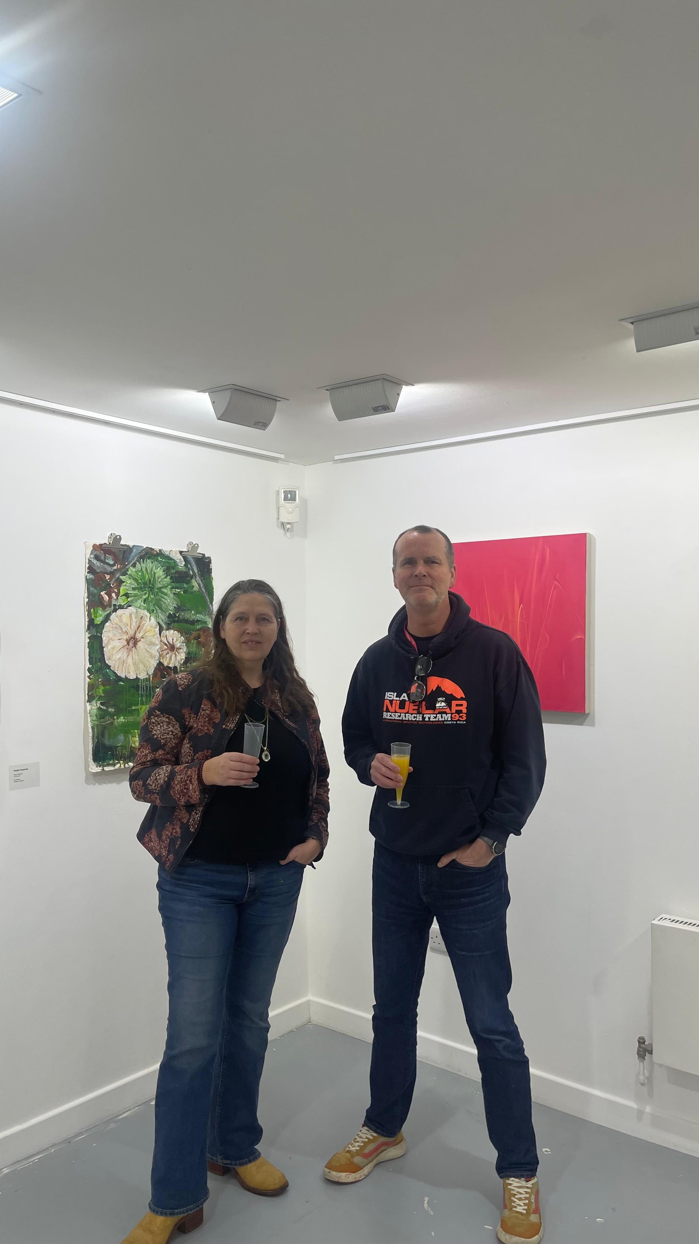 N Karpinski and P Hartley standing in white gallery corner holding drinks, with green botanical artwork and red artwork on walls behind them.