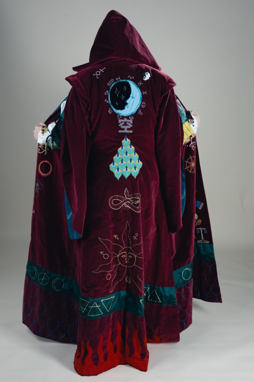 A burgundy hooded robe embroidered with colourful symbols and geometric patterns, shown hanging against a plain grey background.