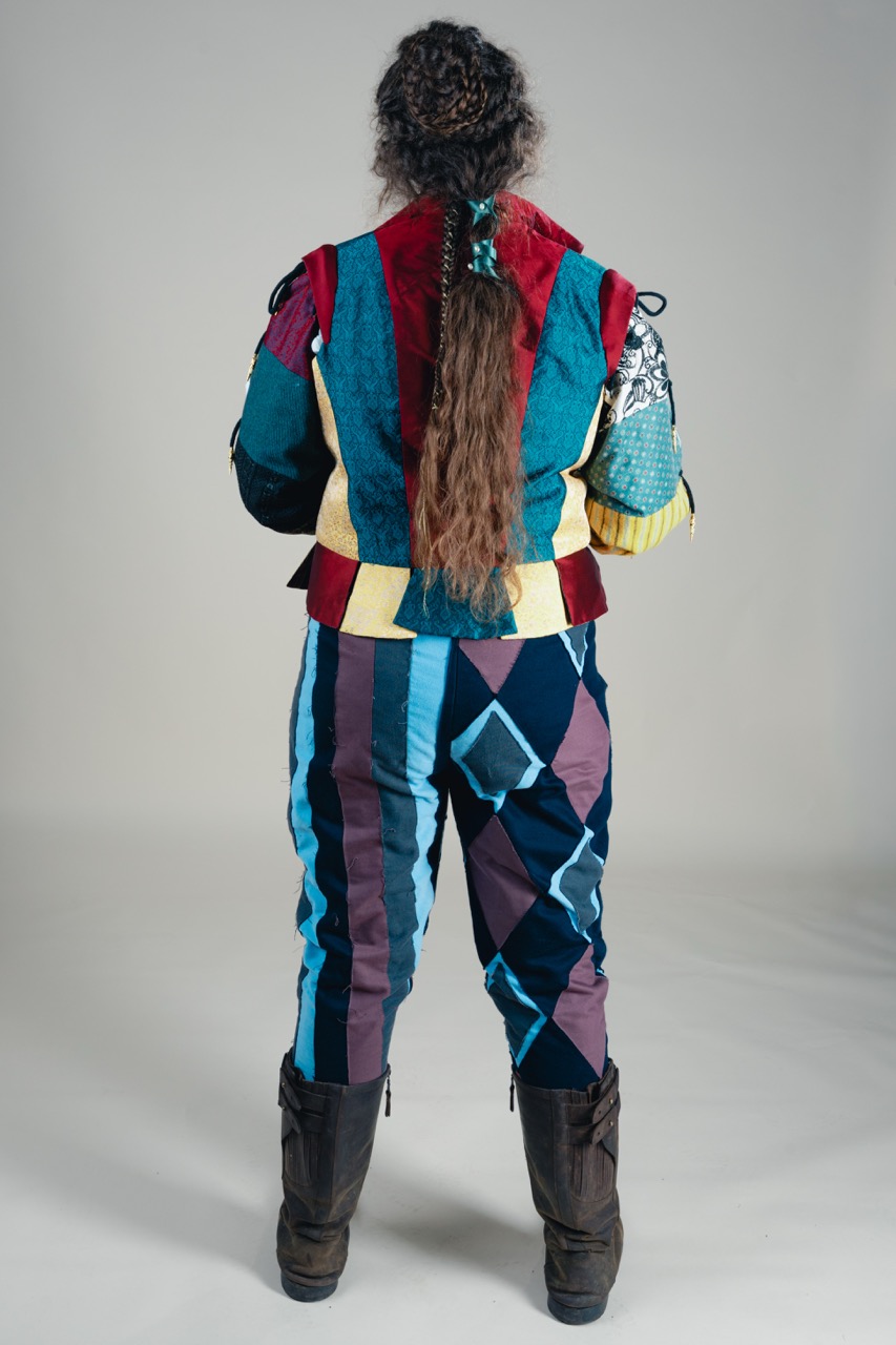 A person viewed from behind wearing a brightly coloured patchwork jacket and trousers, with black boots, standing against a plain grey background.