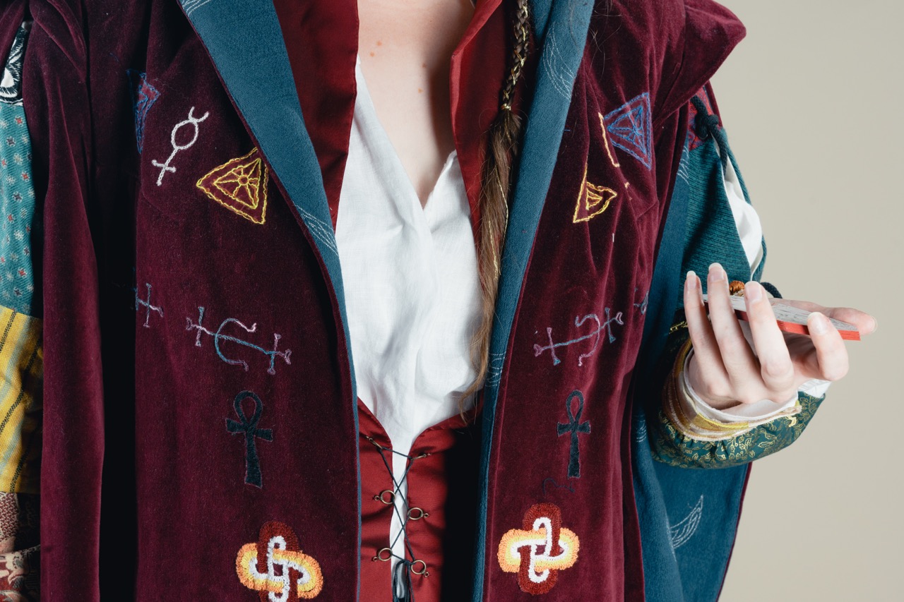 A person wearing a burgundy embroidered vest over a white shirt holds a smartphone, with a beige wall in the background.