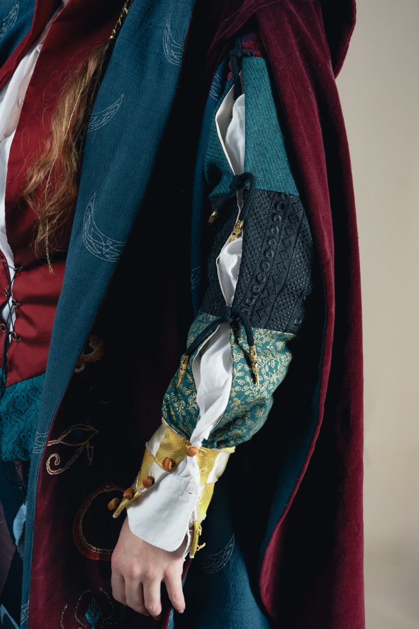 A person wearing layered red and teal clothing with a patterned sleeve and long cape, against a plain grey background.
