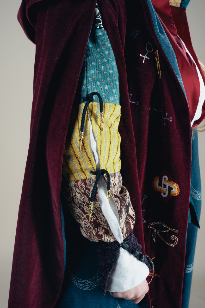 A person wearing a maroon embroidered robe stands with fabric draped over one arm, with patterned yellow and blue sleeves visible underneath.