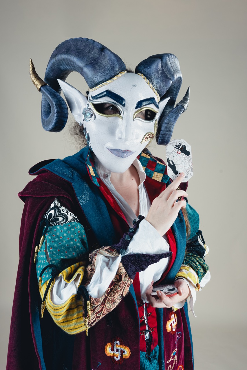 A person wearing a white theatrical mask with curved horns and a colourful costume, holding a small playing card against a plain background.
