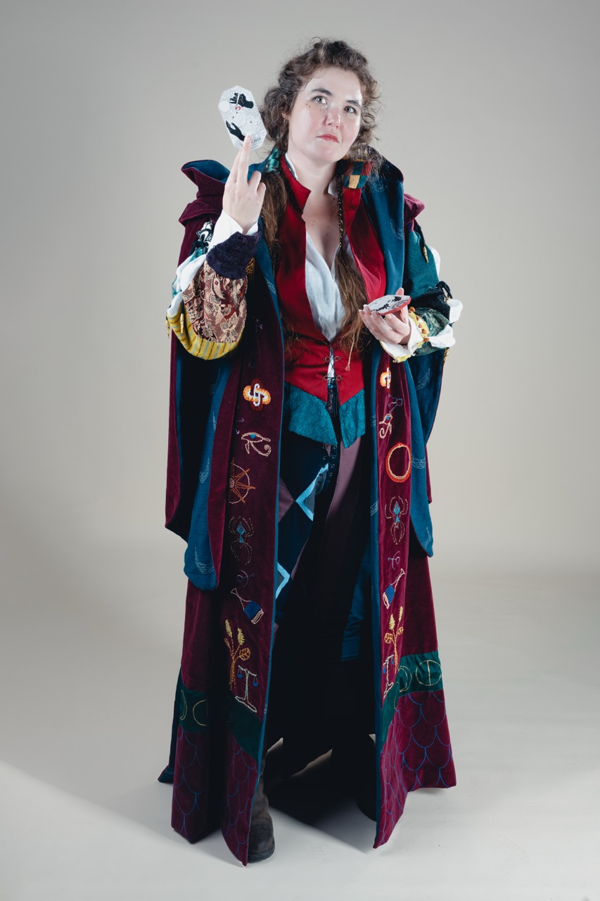 A woman in a long patterned robe and layered clothing stands against a plain grey background, holding a white mask in one hand and a small object in the other.