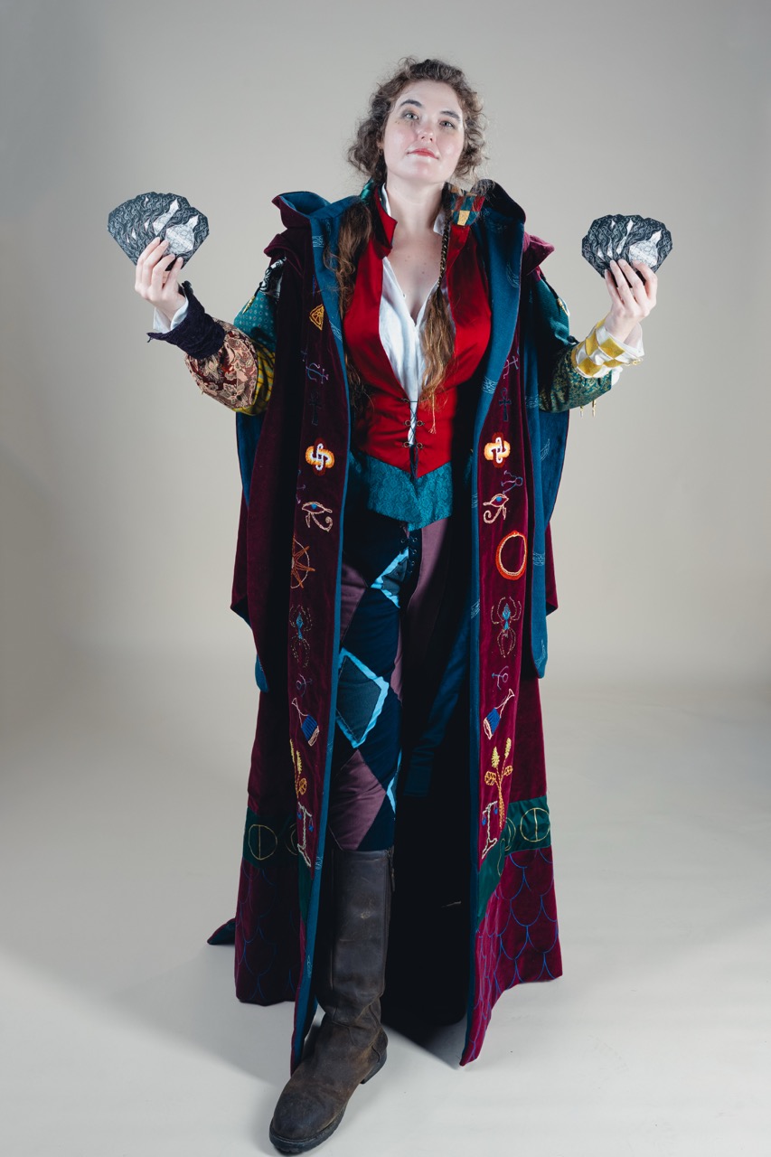 A woman in a long patterned coat and knee-high boots stands against a plain studio backdrop, holding a dark rock in each hand.