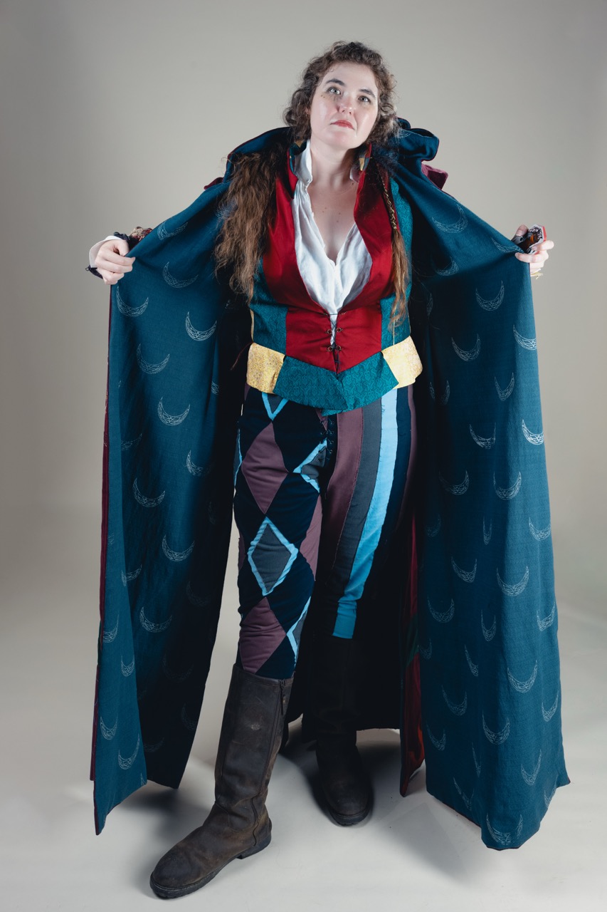 A woman wearing a colourful fantasy costume with a blue crescent-patterned cloak stands against a plain grey background.