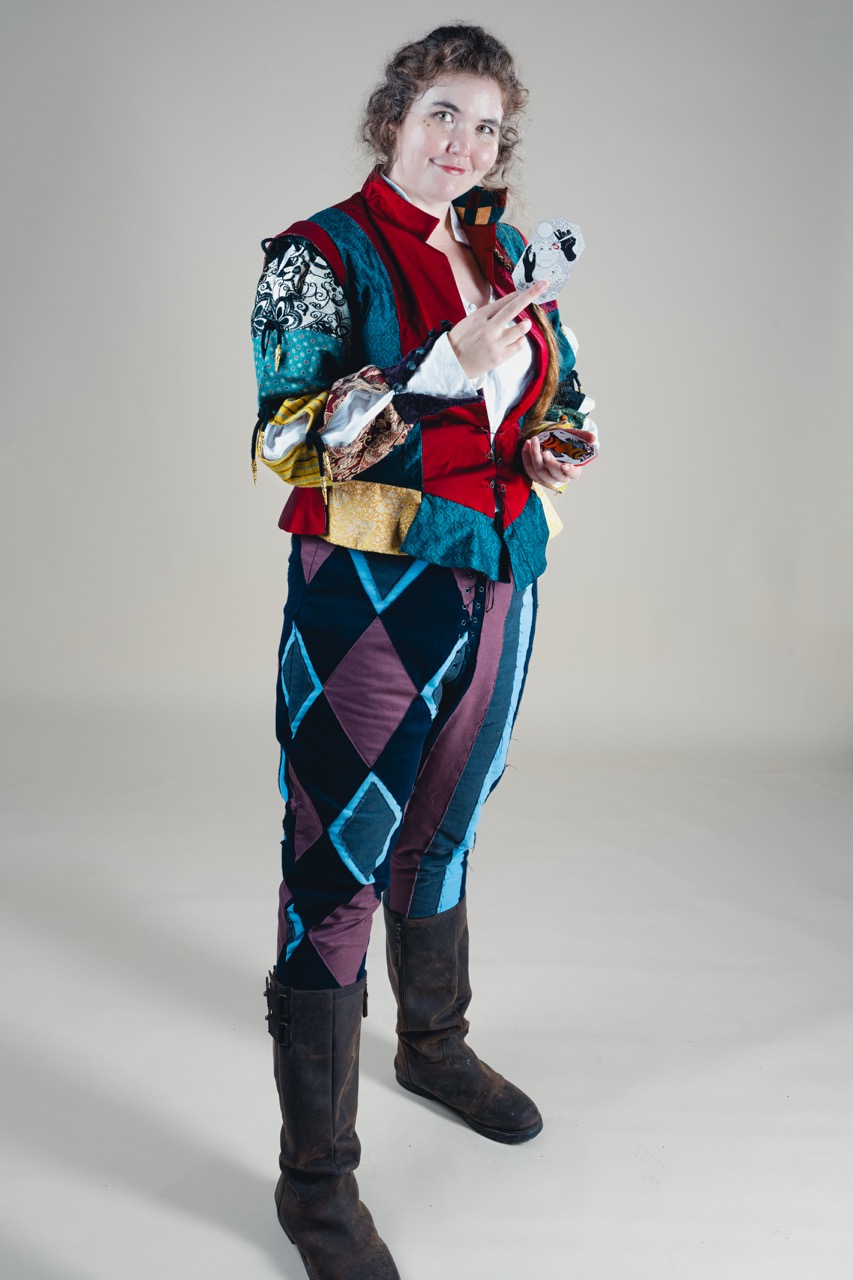 A woman wearing a colourful patchwork costume stands in a studio, holding a small figurine in one hand and a phone in the other.