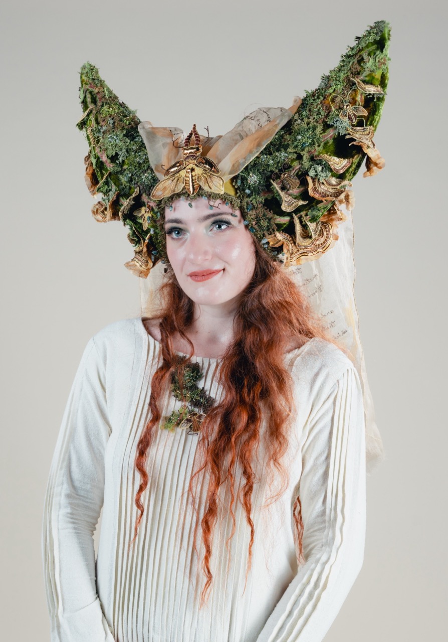 A woman with long red hair wears a white pleated dress and an elaborate moss-covered headdress with butterfly wings against a plain light background.
