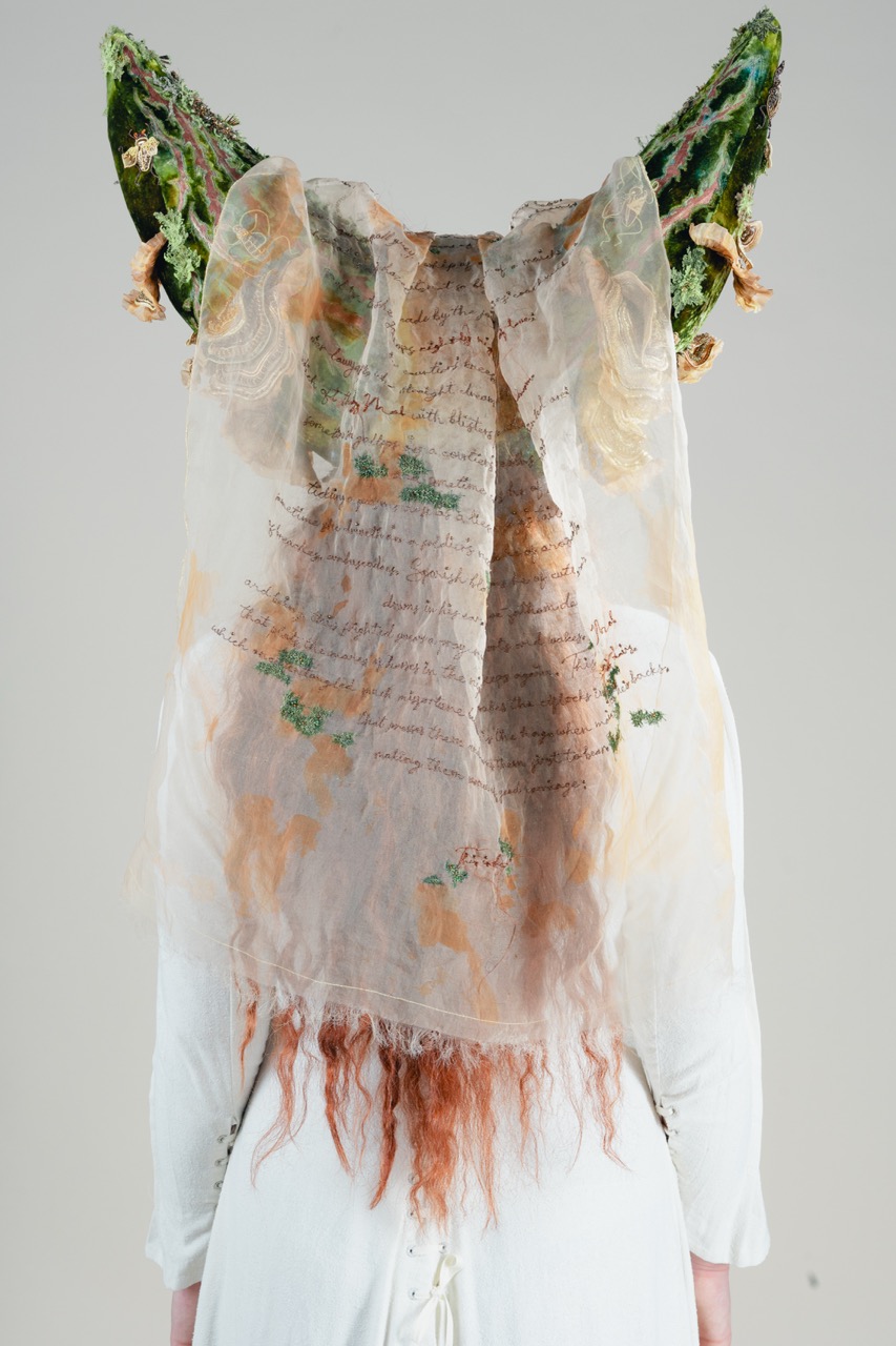 A person wearing a sheer, layered veil with handwritten text and green floral headpiece details against a plain light background.
