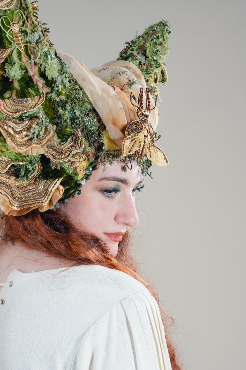 A woman in side profile wearing an elaborate green floral headdress with gold and beige fabric details, against a plain light grey background.