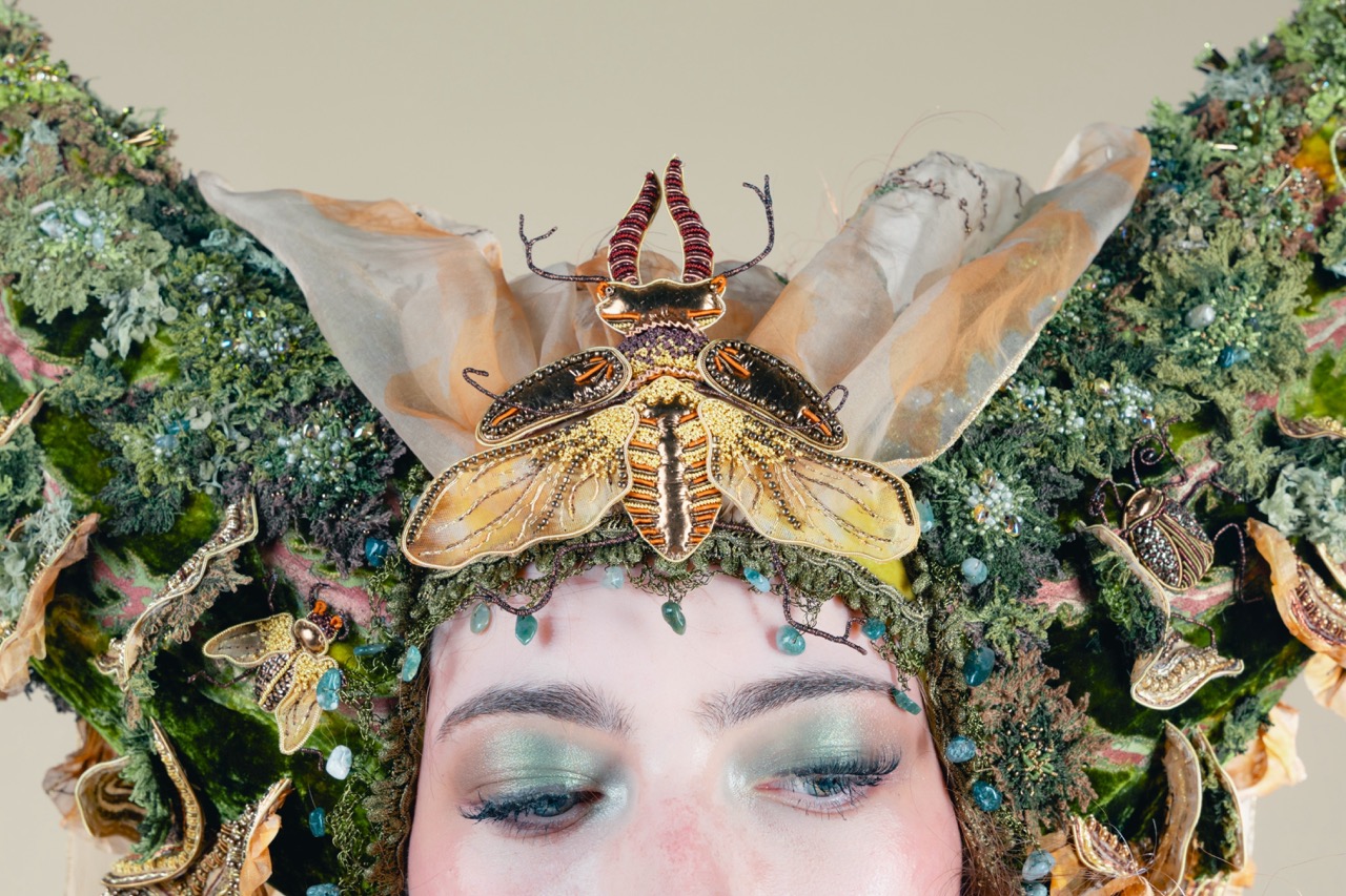 A close-up of a person wearing an ornate headpiece with large moth decorations and green foliage, against a pale background.